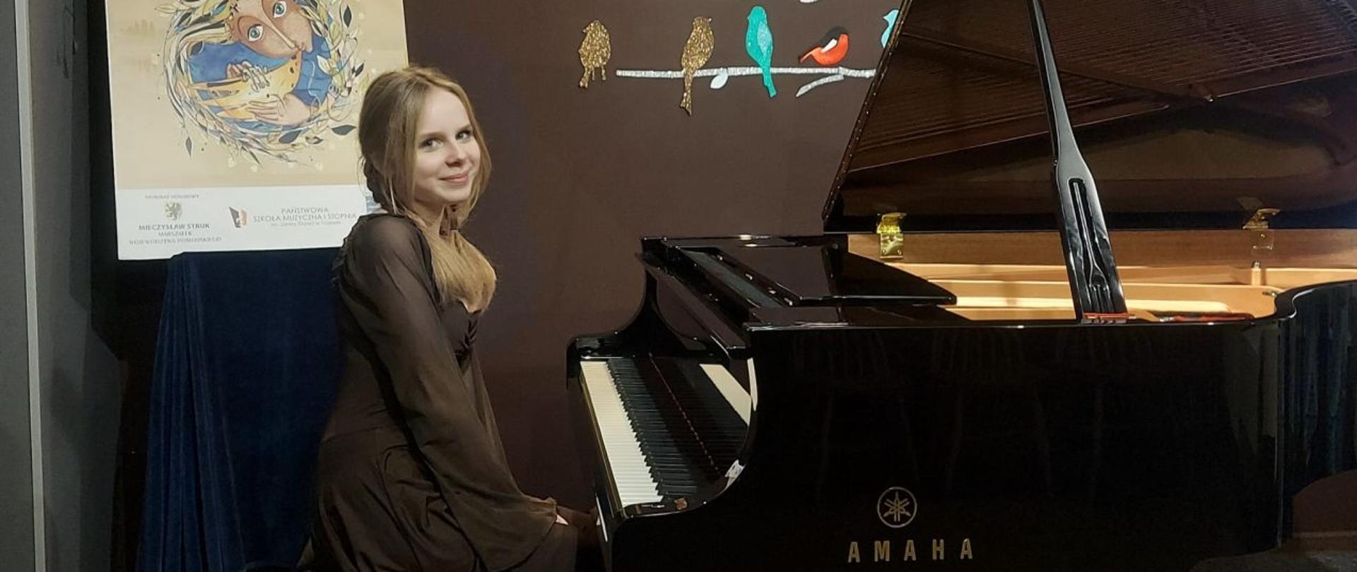 Zdjęcie kolorowe. Dziewczyna o blond włosach ubrana w brązową sukienkę siedzi przy fortepianie 