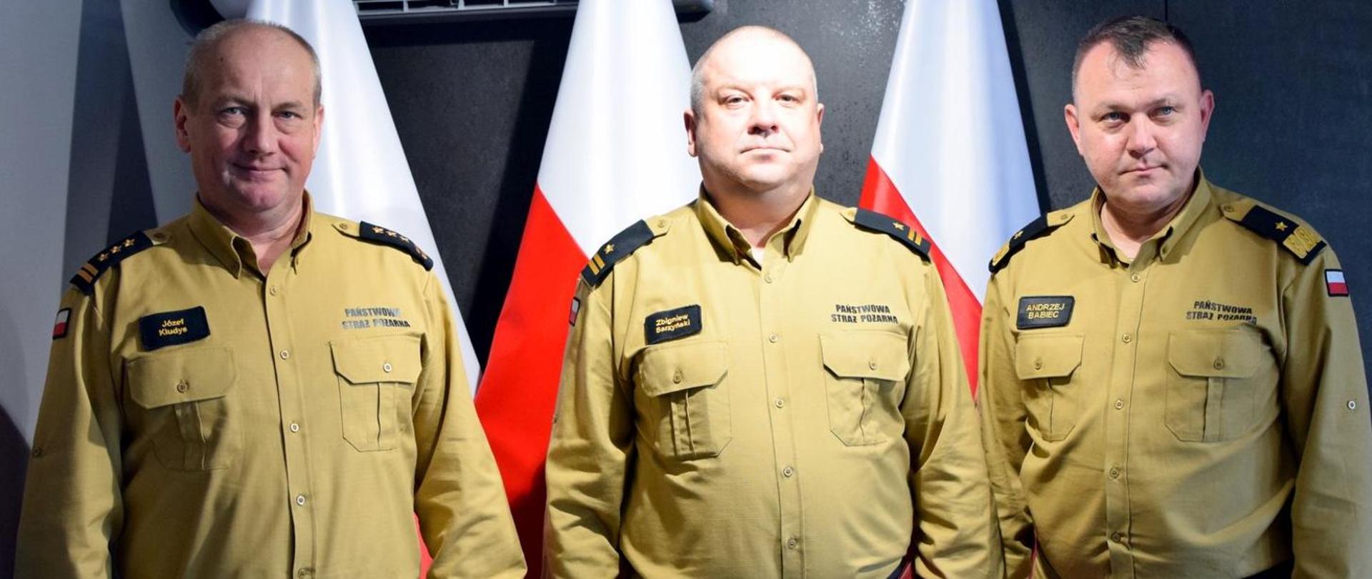 Zdjęcie zrobione wewnątrz pomieszczenia. Na zdjęciu trzech oficerów Państwowej Straży Pożarnej. Za nimi ustawione są biało-czerwone flagi.