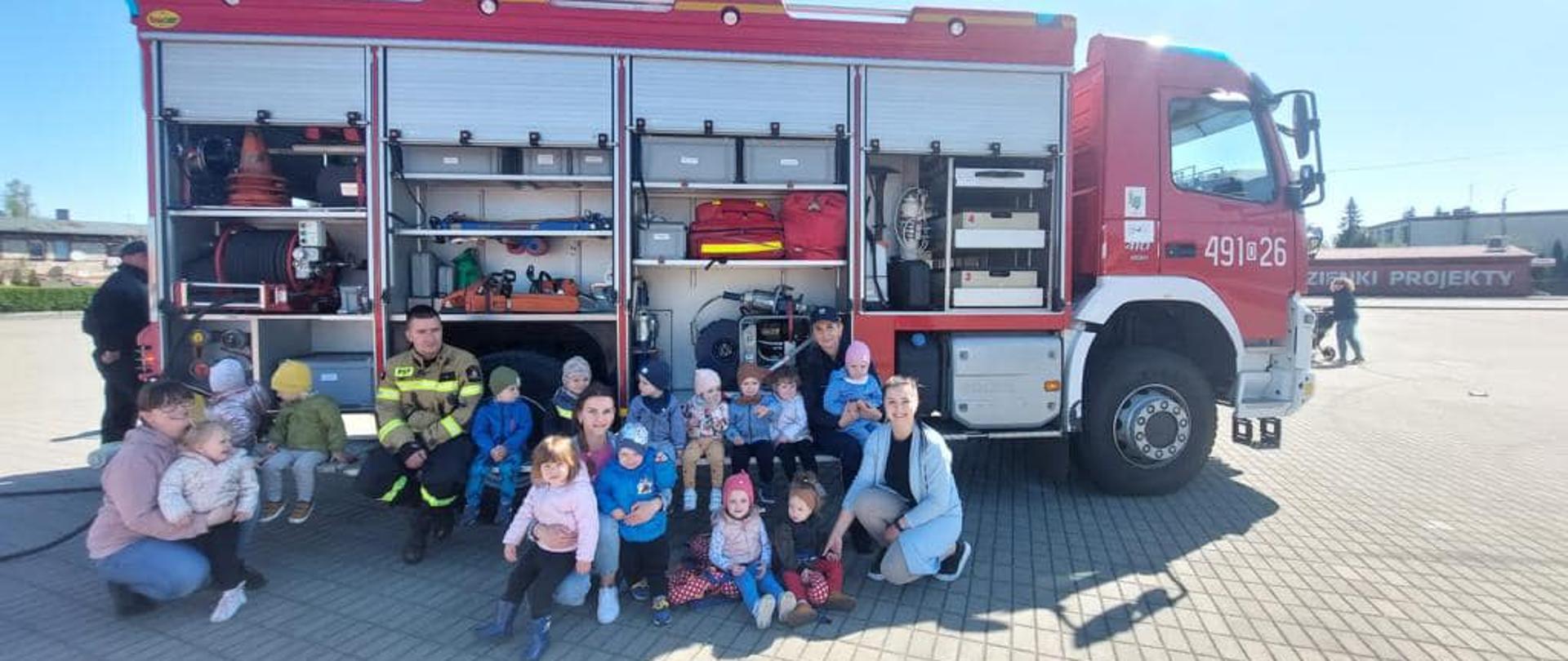 Na zdjęciu znajduje się samochód pożarniczy z otwartymi skrytkami obok przykucają strażacy i opiekunowie oraz dzieci