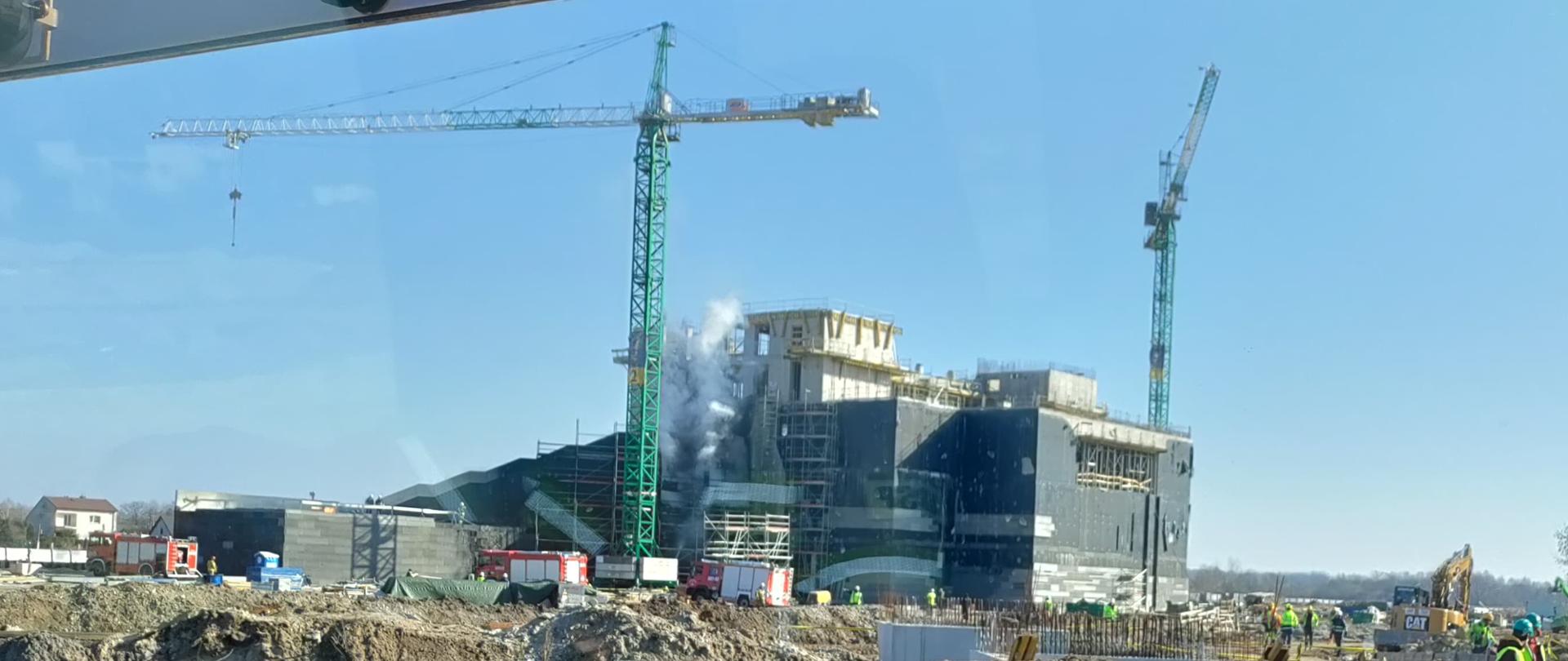 Na głównym planie zdjęcia widoczny jest duży betonowy obiekt przy którym stoją żurawie wieżowe. Przy budynku widoczne są samochody pożarnicze. Z jednej ze ścian wydobywają się kłęby dymu. Po prawej stronie zdjęcia widać grupę robotników budowlanych w jaskrawych strojach oraz kaskach. Zdjęcie wykonane z wnętrza pojazdu strażackiego.