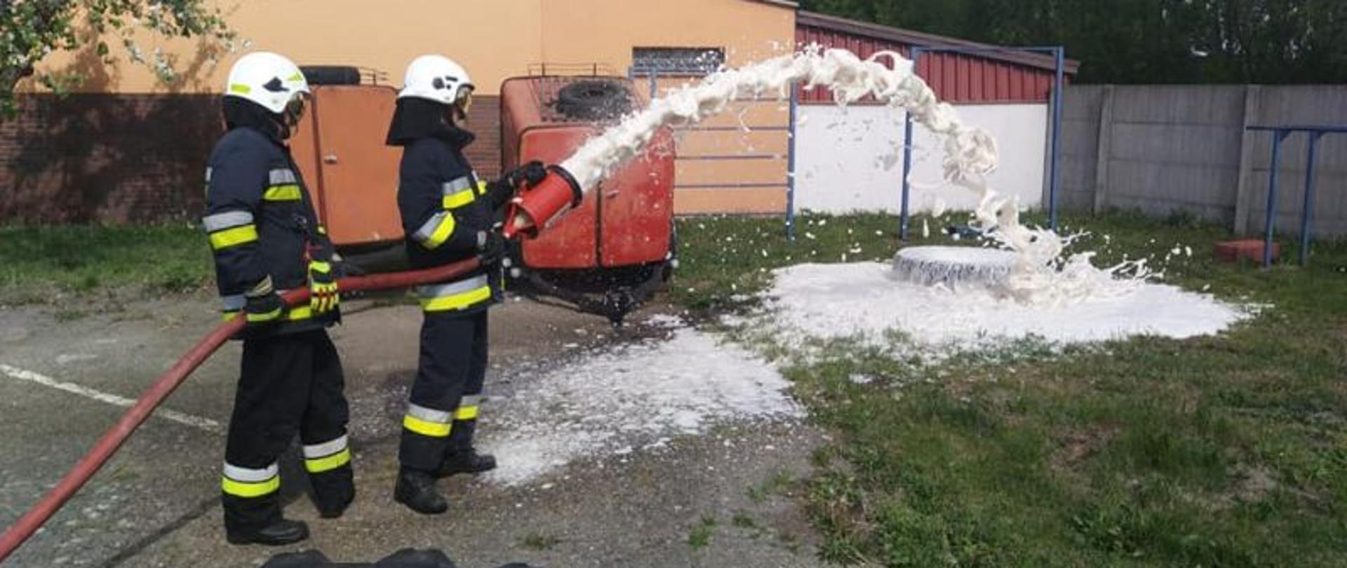 Zdjęcie przedstawia dwóch strażaków. Strażacy mają ciemne mundury, białe hełmy i opuszczone przyłbice. Jeden z nich w rękach trzyma prądownice pianową z której końca leci biała piana.
