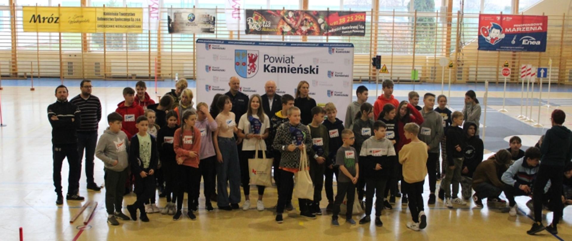 W centralnej części zdjęcia widać grupę młodzieży. Za nimi stoi Starosta kamieński, przedstawiciele Komendy Powiatowej Policji w Kamieniu Pomorskim oraz Powiatowej Stacji Sanitarno-Epidemiologicznej w Kamieniu Pomorskim. Za zgromadzeniem widoczny jest baner z napisem Powiat Kamieński i logo powiatu. 