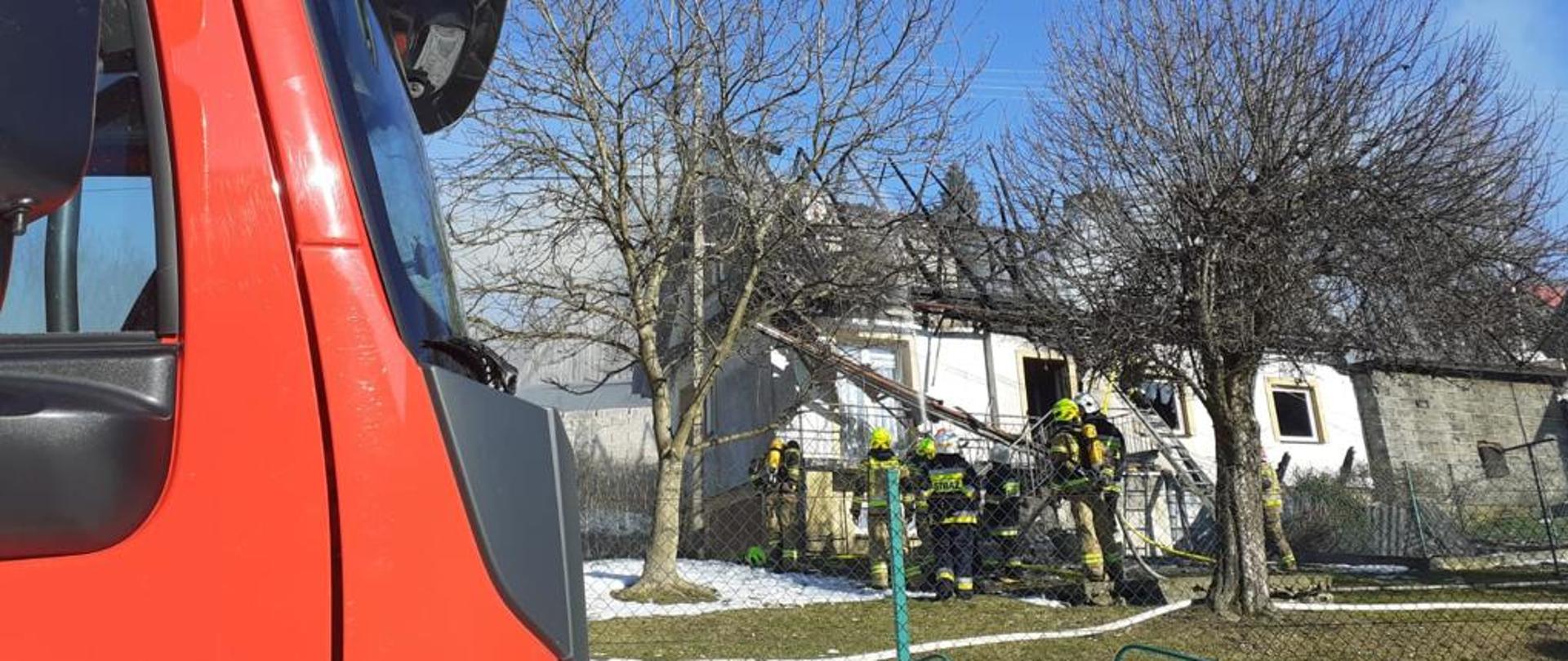 Zdjęcie przedstawia budynek mieszkalny, w którym doszło do pożaru. Widoczna spalona konstrukcja dachu, wiszące elementy konstrukcyjne. Przed obiektem widoczna jest grupa strażaków. Nad obiektem unosi się delikatny jasny dym.