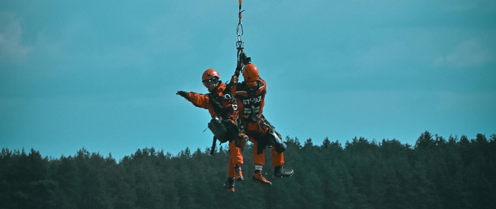 Dwóch ratowników PSP w czerwonych kombinezonach podwieszonych na linie pod śmigłowcem, wykonujących ćwiczenia wysokościowe nad polaną.