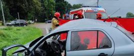 Fotografia przedstawia powypadkowy wrak pojazdu oraz strażaków podczas prowadzenia działań ratowniczych.