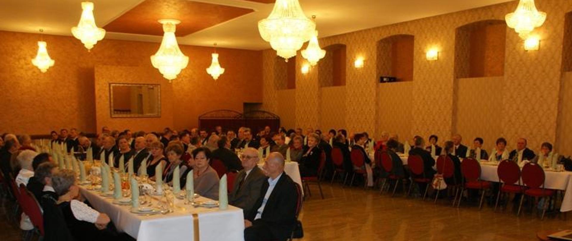 Na zdjęciu widzimy kilkadziesiąt osób mężczyzn i kobiet siedzących przy stole. Stół nakryty jest białym obrusem we wzory, na stole widzimy talerze, szklanki, napoje oraz serwetki. Osoby znajdują się w pomieszczeniu, dalej widzimy wystrój tego wnętrza, ściany pokryte żółtą tapetą we wzorki. Na ścianie widzimy też świecące kinkiety. Z sufitu zwisają brylantowe żyrandole. Podłoga parkiet koloru żółtego.