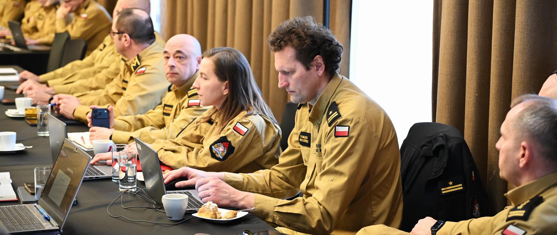 Za długim stołem, z czarnym suknem, siedzą strażacy, w tym również kobiety. Wszyscy w służbowym, piaskowym mundurze. Na stole mają naczynia z napojami, ciasto. Strażak na pierwszym planie pracuje z laptopem. 