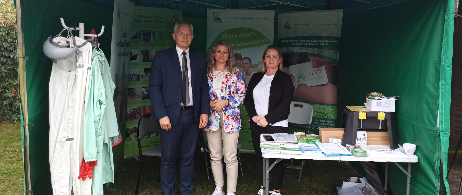 Mężczyzna i dwie kobiety pod zielonym namiotem KRUS z napisem „Pracuj bezpiecznie”