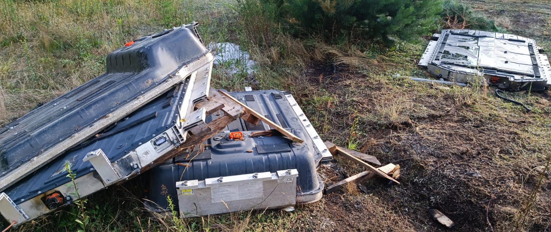 Zdjęcie przedstawia porzucone na łące duże akumulatory do samochodów elektrycznych. Trzy baterie lezą na suchej trawie. W tle widać niewielkie sosny.