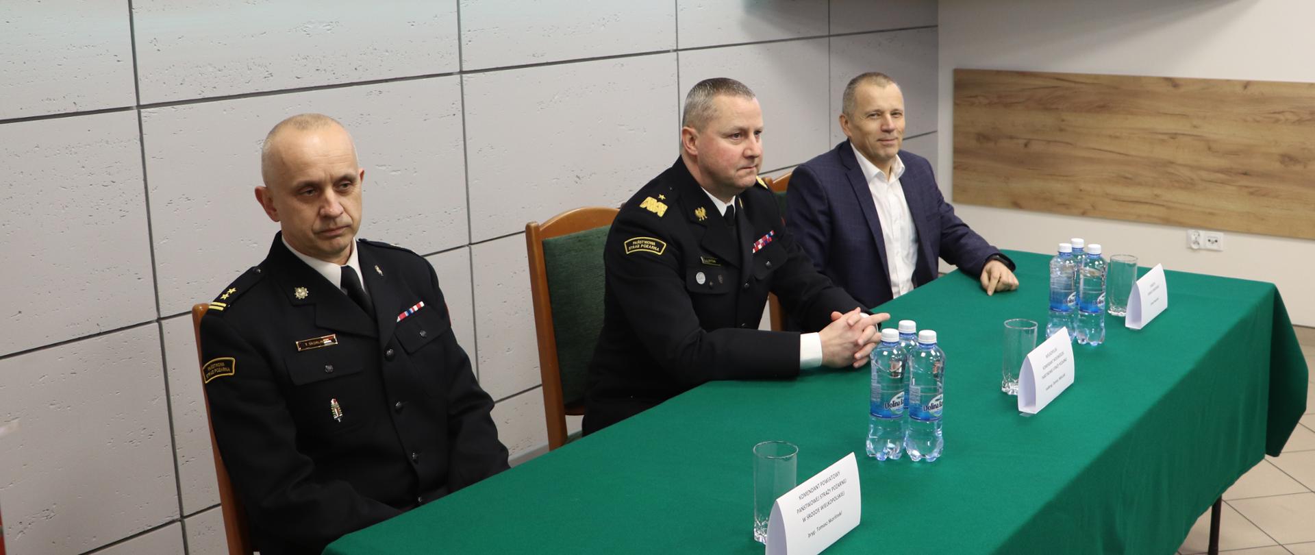 trzech mężczyzn siedzi przy stole nakrytym zielonym obrusem, na stole butelki z wodą w tle ekran z prezentacją