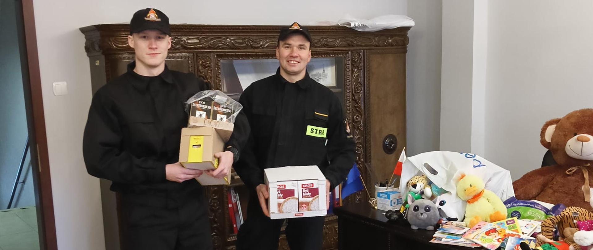 Dwóch strażaków przekazuje dary, które zostały zebrane w ramach akcji dla dzieci i seniorów. 