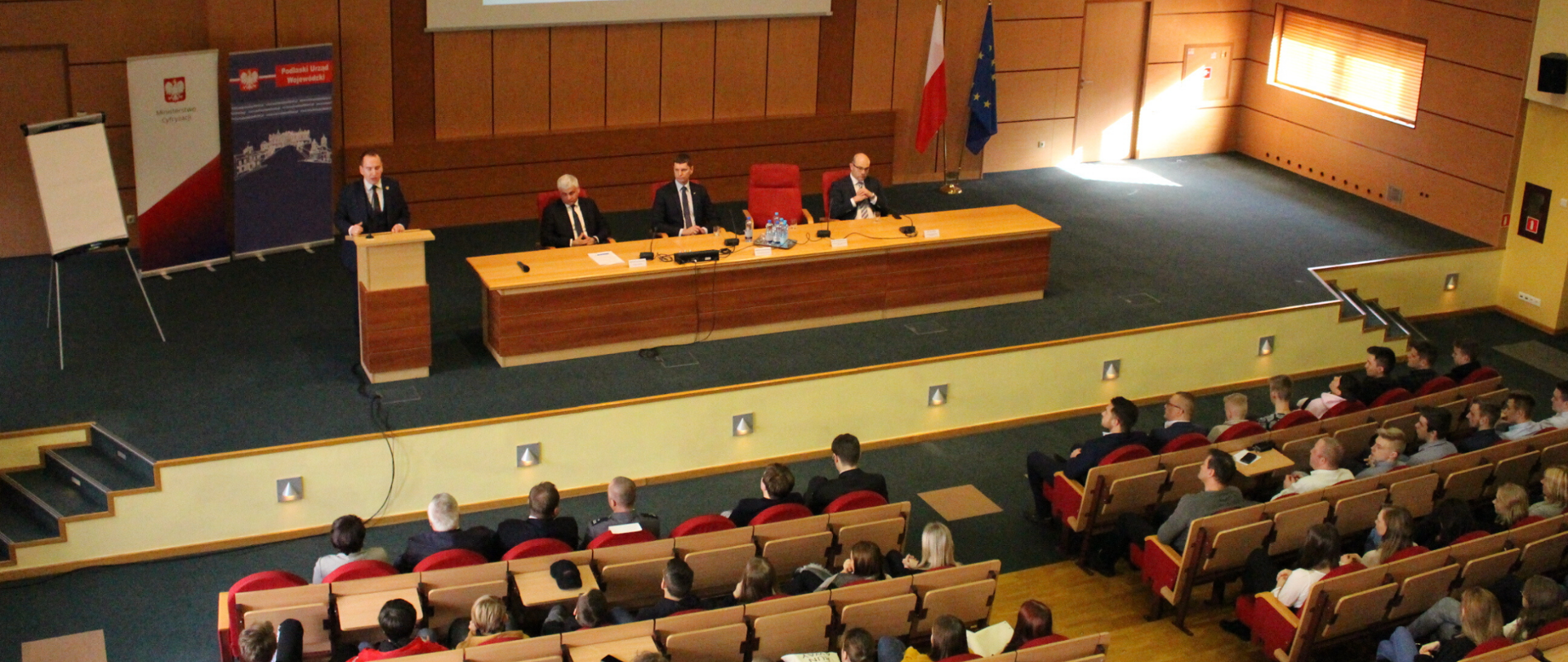 Sala w Podlaskim Urzędzie Wojewódzkim. Przy mównicy stoi minister Adam Andruszkiewicz. Obok przy stole siedzą m.in. wojewoda podlaski i minister edukacji. Przed ministrem siedzą słuchacze, przeważają młode osoby.