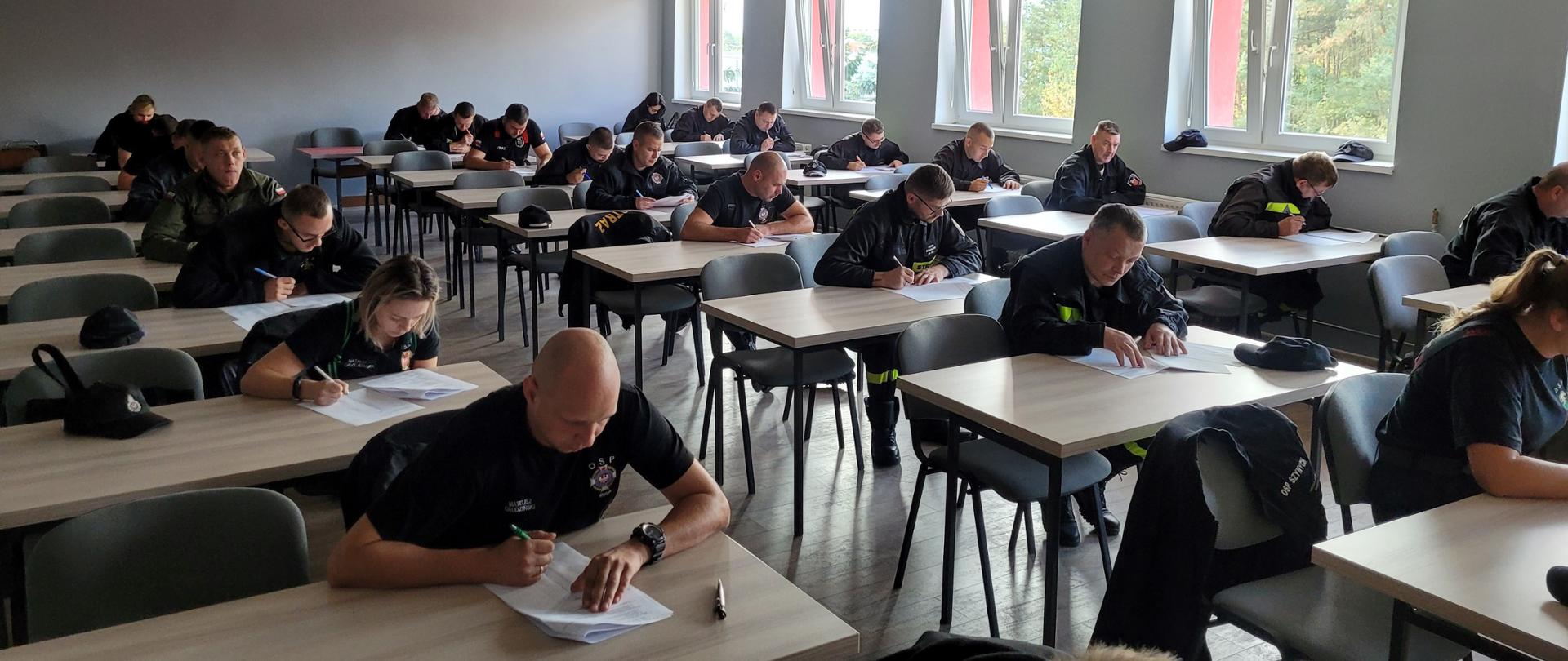 Sala wykładowa z oknami widocznymi po prawej stronie, w której pojedynczo przy stołach siedzą strażacy w czarnych mundurach i wypełniają pisemny test.