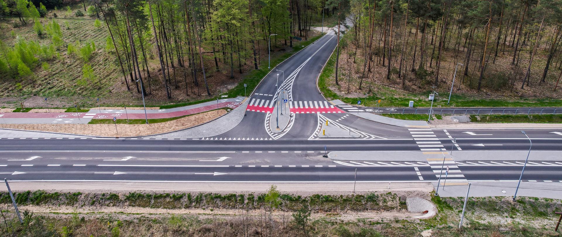 Zdjęcie przedstawia drogę krajową DK46, biegnącą przez malowniczy krajobraz. Droga jest asfaltowa, jednopasmowa w każdym kierunku, z wyraźnie oznaczonymi pasami ruchu. Po jej bokach znajdują się ścieżki rowerowe, oddzielone pasem zieleni. Wzdłuż drogi widoczne są znaki drogowe oraz elementy infrastruktury, takie jak barierki ochronne i oświetlenie uliczne. Otaczający teren składa się z pól, lasów oraz pojedynczych zabudowań. W tle widać łagodne wzgórza, które dodają krajobrazowi głębi i harmonii. Zdjęcie ukazuje dobrze zaprojektowaną trasę, łączącą nowoczesną infrastrukturę z naturalnym otoczeniem.