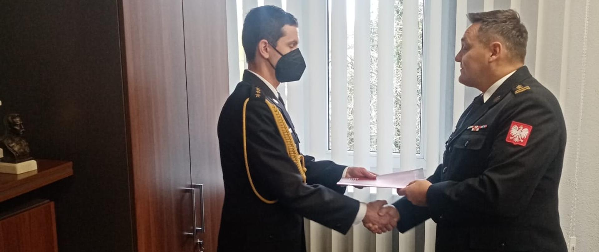 Na zdjęciu strażacy ubrani w mundurach wyjściowych stoją w pomieszczeniu biurowym, witają się przez podanie prawej dłoni i przekazują dokumenty papierowe.