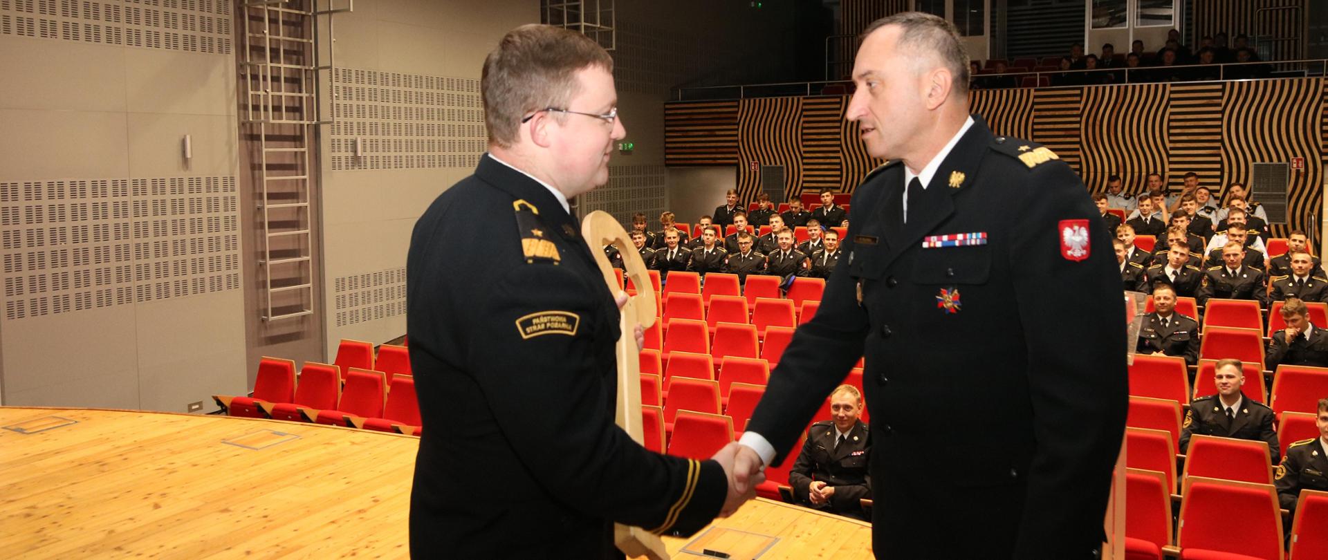 Nadbryg. Piotr Filipek na scenie szkolnej Auli wraz z nim przyjmujący symboliczny klucz do Szkoły przewodniczący Rady Kadetów kdt Michał Ziętarski. Mężczyźni ściskają swoje prawe dłonie. 