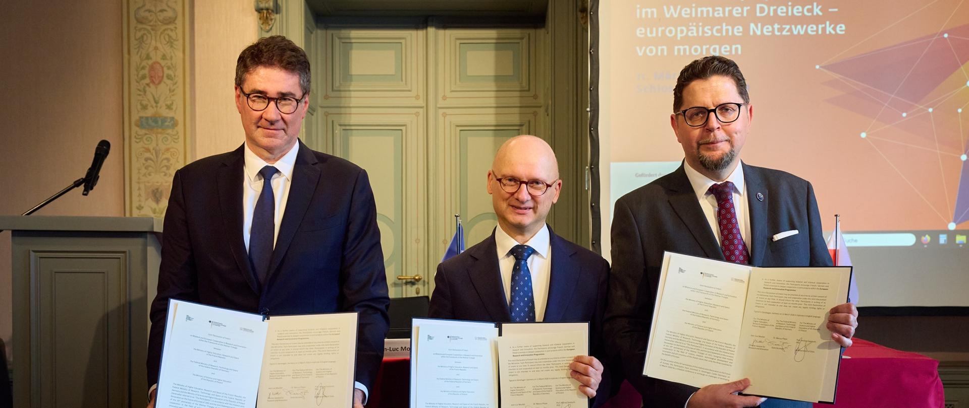 Przed nakrytym czerwonym materiałem stołem stoi trzech mężczyzn w garniturach, pokazują trzymane w rękach otwarte teczki z dokumentami.