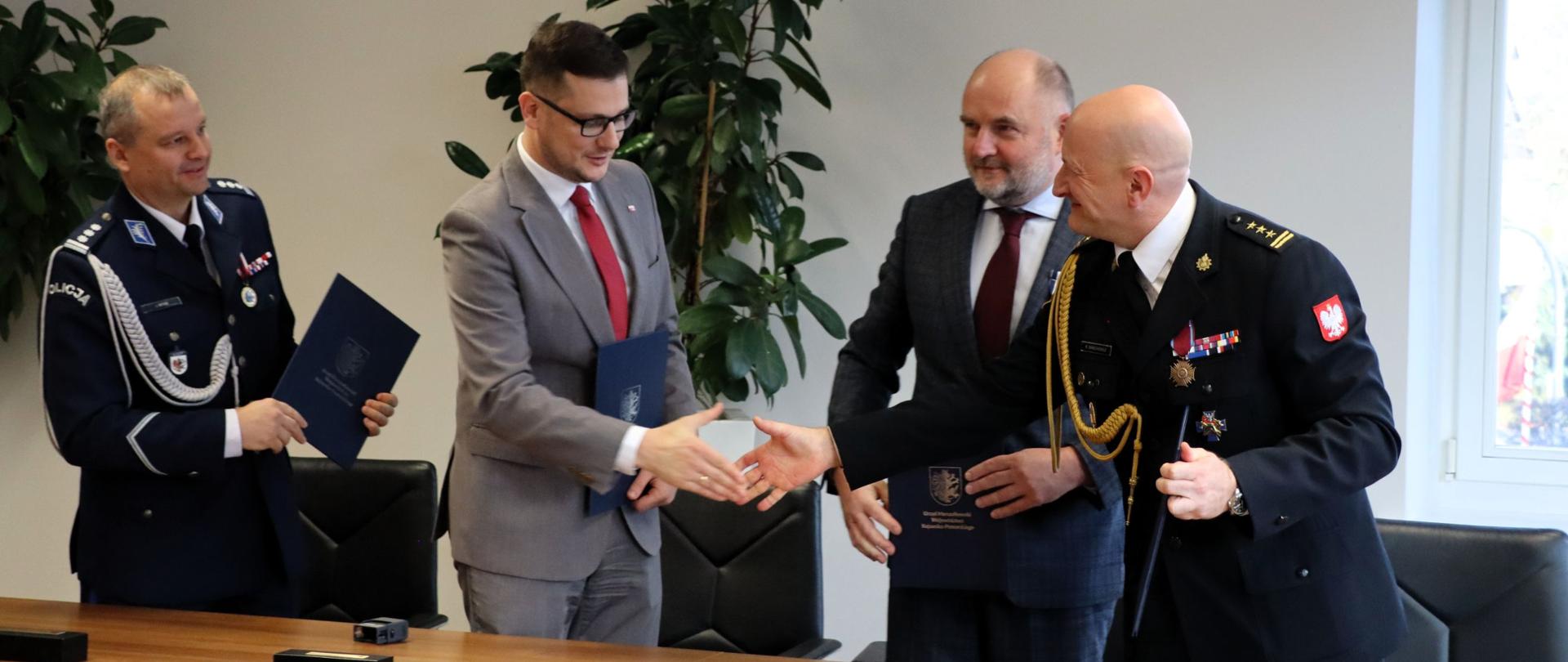 Za stołem, na stojąco policjant, wojewoda, marszałek i strażak. Wymieniają uściski dłoni. Na ścianie herb województwa, stojące rośliny ozdobne jako wystrój wnętrza. 