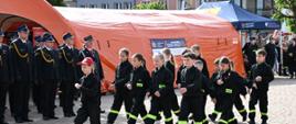 Zdjęcie przedstawia grupę dzieci z Młodzieżowej Drużyny Pożarniczej maszerującą po wybrukowanym placu podczas uroczystości Święta Ogniowego. Dzieci ubrane są w czarne mundury koszarowe z odblaskowymi, żółtymi pasami na nogawkach i napisem „STRAŻ” na plecach lub piersi. Na czele maszeruje dziewczynka w czerwonej czapce z daszkiem. W tle, po lewej stronie, stoją wyprostowani strażacy w ciemnych mundurach galowych, a za nimi widoczny jest duży pomarańczowy namiot pneumatyczny z logotypami Akademii Ochrony Ludności i Obrony Cywilnej.
