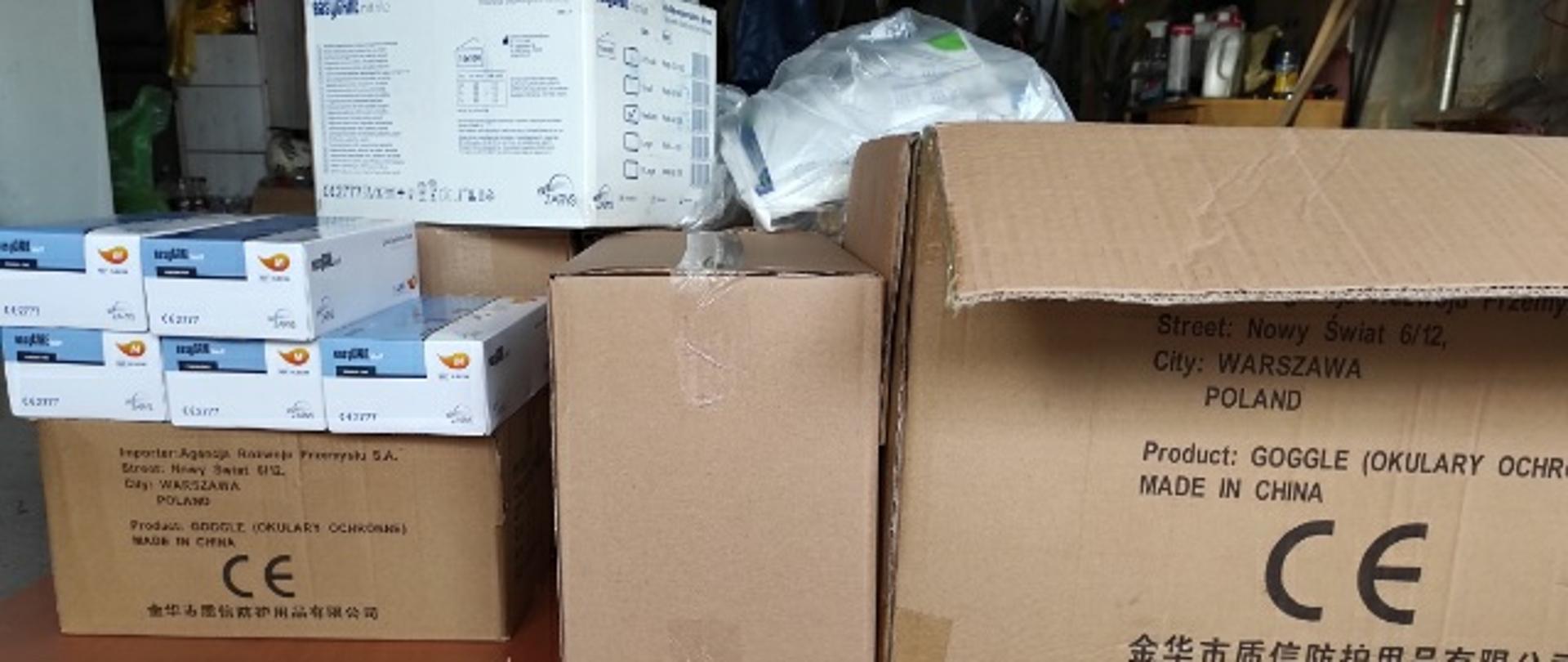 W garażu stoją kartony, na których leżą pudełka rękawiczek lateksowych i maseczki. 