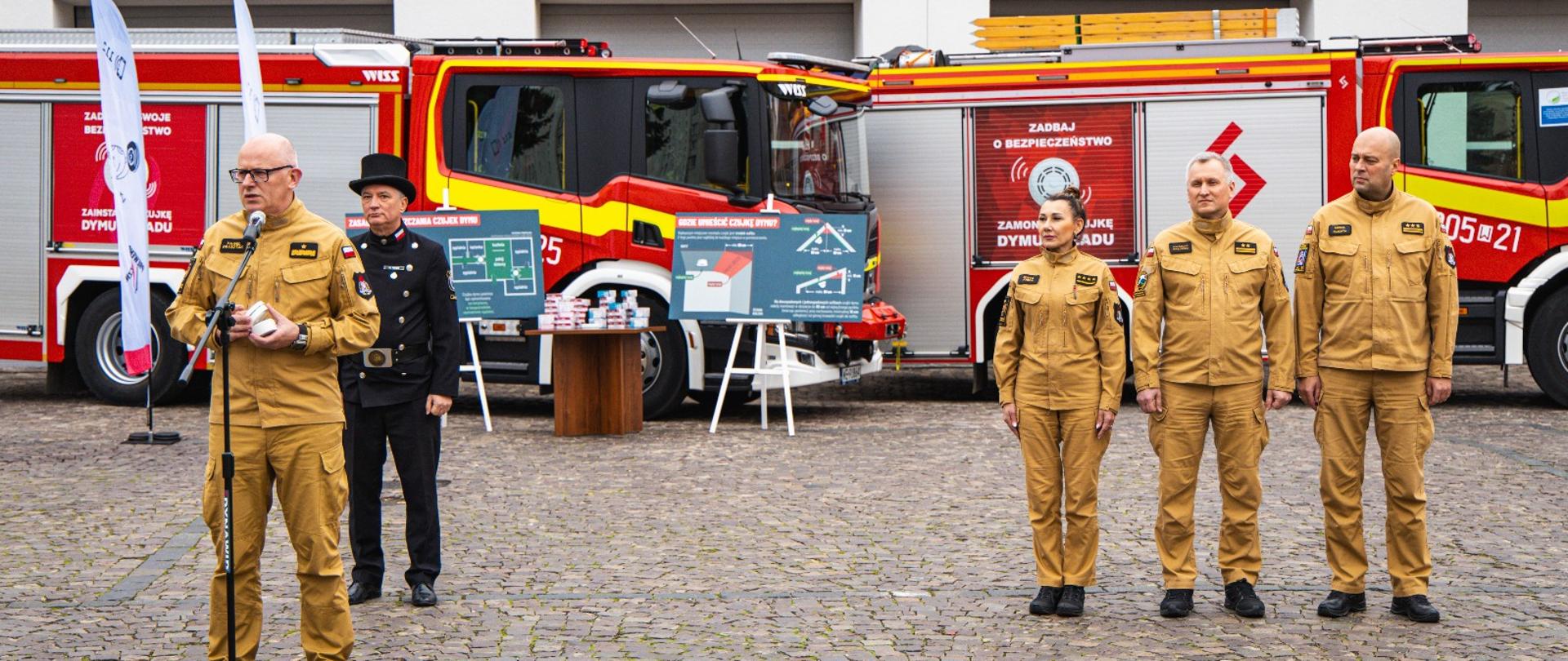 Pięć osób w mundurach stoi na brukowanym placu przed budynkiem. Po lewej stronie mężczyzna w piaskowym kombinezonie z emblematami służb ratowniczych przemawia do mikrofonu na statywie. Za nim stoi mężczyzna w czarnym płaszczu i meloniku. Po prawej stronie w linii stoją kobieta i dwóch mężczyzn, również w piaskowych kombinezonach. W tle widać dwa czerwono-żółte wozy strażackie z polskimi napisami dotyczącymi bezpieczeństwa, a także stojaki z tablicami informacyjnymi.