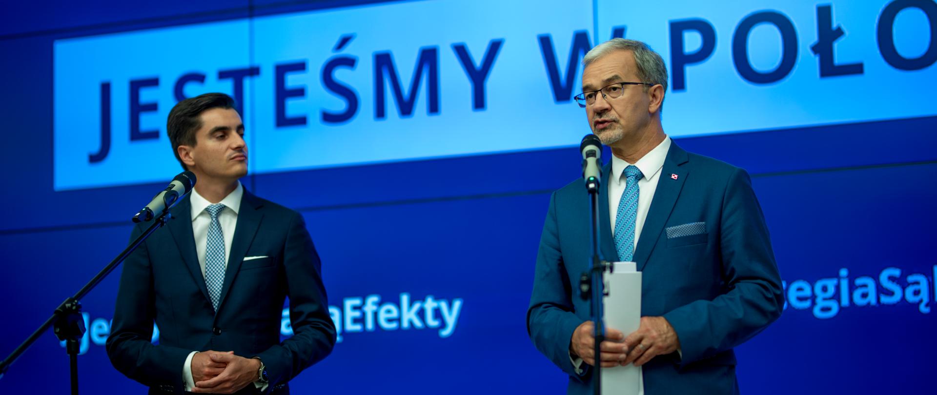 Zdjęcie z konferencji prasowej. Na pierwszym planie dwie osoby stojące przed mikrofonami na statywach. Od lewej Tomasz Janka, dyrektor Rządowego Biura Monitorowania Projektów i minister inwestycji i rozwoju Jerzy Kwieciński. Za nimi ekran z uciętym napisem "Strategia na rzecz Odpowiedzialnego Rozwoju. Jesteśmy w połowie drogi". 