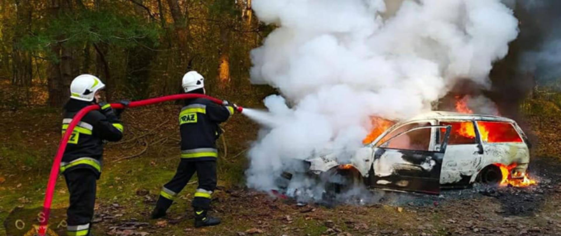Dwóch strażaków ochotników gasi jednym prądem wody palący się w całości samochód osobowy, zaparkowany na leśnej drodze.