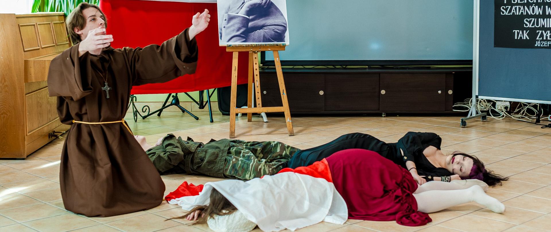 Zdjęcie przedstawia inscenizację patriotyczną w pomieszczeniu z okazji Święta Niepodległości (11 listopada).
Centralny element: sztaluga z czarno-białym portretem historycznej postaci Józefa Piłsudskiego, obok polska flaga z godłem.
Osoby na scenie: jedna osoba w brązowym habicie, z rozłożonymi rękami w geście modlitwy, trzy osoby leżące na podłodze – jedna w ubraniu maskującym (wojskowym), druga w czerwono-białym stroju, trzecia w czarnej długiej sukni. Tło: tablica z cytatem, duża roślina doniczkowa, ekran projekcyjny.
Całość jest scenką historyczno-patriotyczną, symbolizującą walkę o wolność i ofiarę dla ojczyzny.