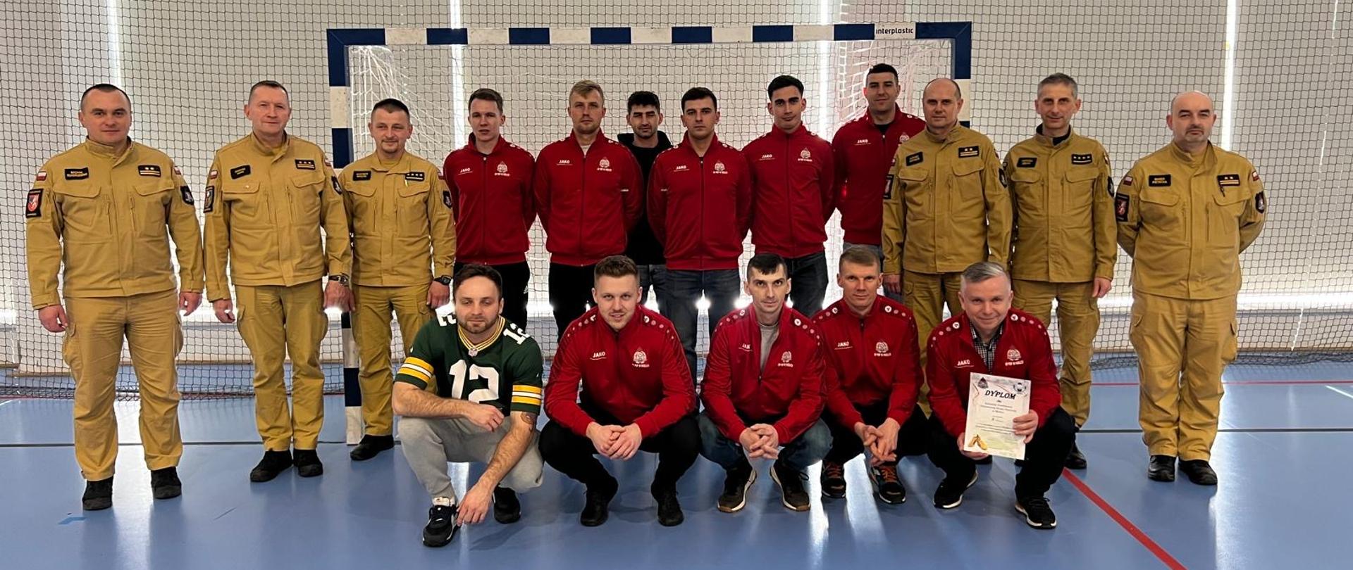 Grupa siedemnastu mężczyzn pozuje na hali sportowej przed bramką do gry w futsal podczas turnieju eliminacyjnego XXVI MISTRZOST WOJEWÓDZTWA PODKARPACKIEGO STRAŻAKÓW PSP w Futsalu. Część osób ubrana jest w beżowe mundury strażackie z czarnymi naszywkami i oznaczeniami, a część w czerwone dresy sportowe z herbem PSP. W pierwszym rzędzie, klęczy oraz przykuca pięciu mężczyzn – czterech w czerwonych dresach i jeden w zielonej koszulce sportowej z numerem „12”. Jeden z nich trzyma w dłoni dyplom. Hala ma niebieską podłogę z żółtymi i czerwonymi liniami, a za grupą widoczna jest siatka ochronna oraz duże okna wpuszczające naturalne światło. Wszyscy patrzą w stronę aparatu, a niektórzy uśmiechają się.