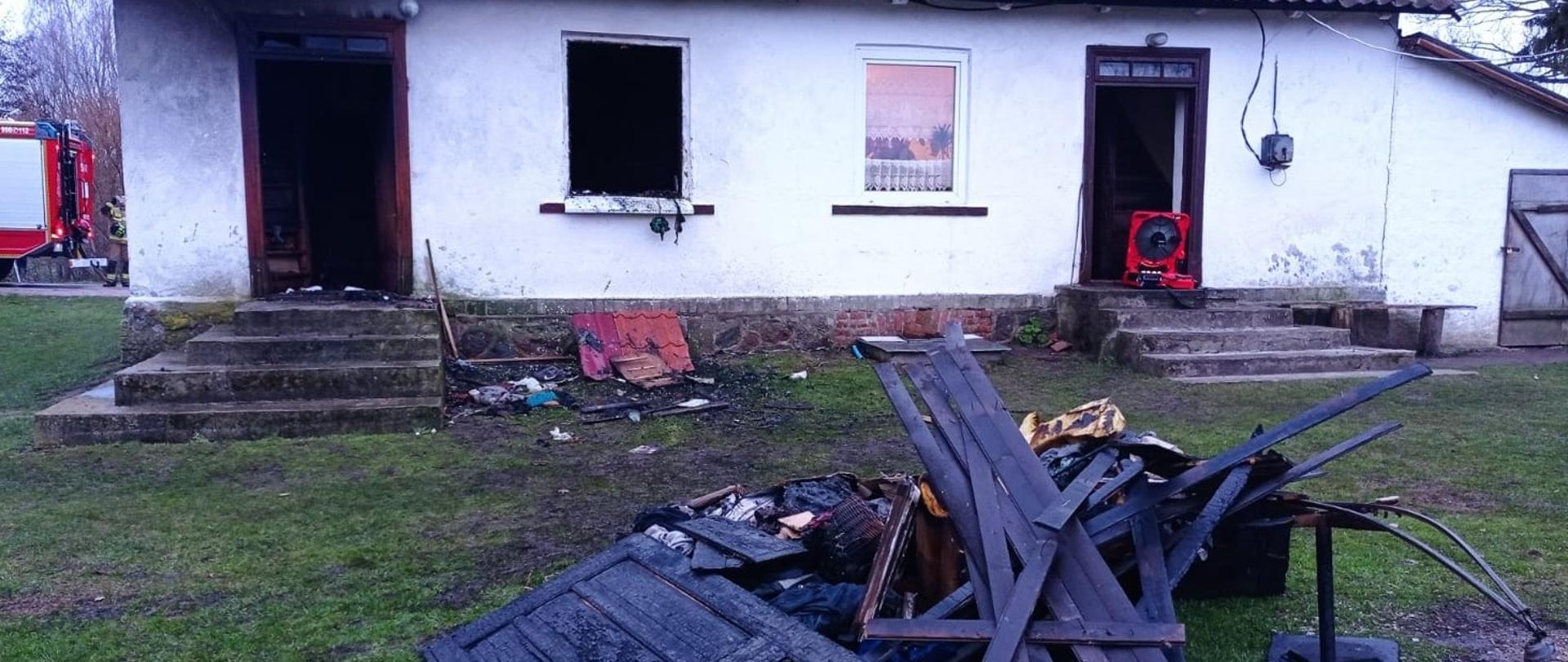 Budynek jednorodzinny biały, otwarte dwa wejścia do budynku i jedno okno. Przed budynkiem widać wyniesione spalone elementy wyposażenia. Po lewej stronie widoczny jest samochód gaśniczy przy, którym stoi strażak.