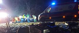 Strażacy działają z wykorzystaniem sprzętu ratowniczego przy uszkodzonych samochodach na jezdni. Zdjęcie wykonane w porze nocnej.