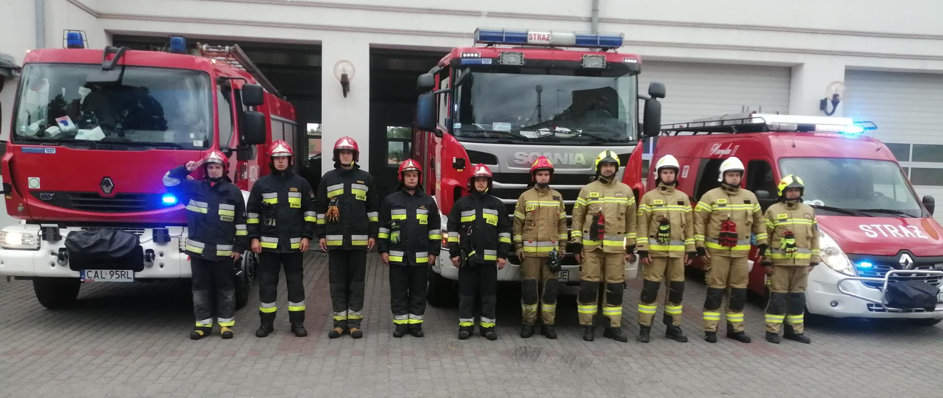 Zdjęcie obrazuje stojących w szeregu dziesięciu funkcjonariuszy. Pięciu funkcjonariuszy z Państwowej Straży Pożarnej stoją w umundurowaniu specjalnym koloru granatowego z żółto-srebrnymi emblematami odblaskowymi oraz założonymi na głowach hełmami w kolorze czerwonym. Zaraz obok stoją strażacy ochotnicy w umundurowaniu specjalnym koloru piaskowego z założonymi hełmami na głowach koloru białego. Strażacy stoją na tle trzech pojazdów pożarniczych w kolorze czerwono-białym. Samochody zostały wyprowadzone przez otwarte bramy garażowe z budynku jednostki ratowniczo-gaśniczej, która jest w kolorze białym. Samochody wysyłają sygnały świetlne w kolorze niebieskim (koguty).