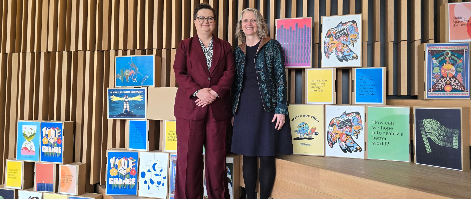 Na tle prostokątów z materiałami edukacyjnymi do zdjęcia pozują 2 kobiety, w tym wiceminister Katarzyna Lubnauer ubrana w czerwony garnitur i białą koszulę z czarną kratą. Postacie stoją na schodach, w nowoczesnej przestrzeni.