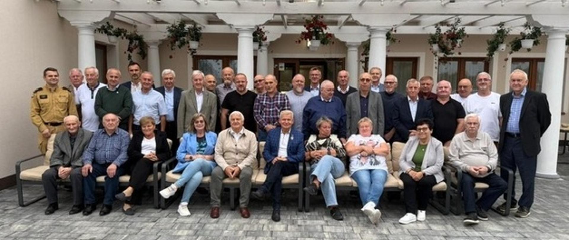 Uczestnicy spotkania pozują siedząc i stoją przed budynkiem. 