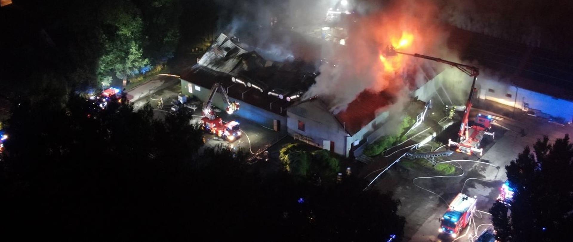 Noc, zdjęcie zrobione z dronem. Czerwone strażackie wozy bojowe, widziane z góry mają wyciągnięte do góry ramiona z koszami, wewnątrz są strażacy ubrani w mundury bojowe, na głowach mają czerwone hełmy. Cały teren jest oświetlony. Budynki częściowo objęte pożarem, zawalony dach na jednym z obiektów. Podawana jest woda na palące się obiekty. Nad obiektami unoszą się kłęby czarnego dymu. Częściowo widać oświetlone drzewa z lewej strony zdjęcia.