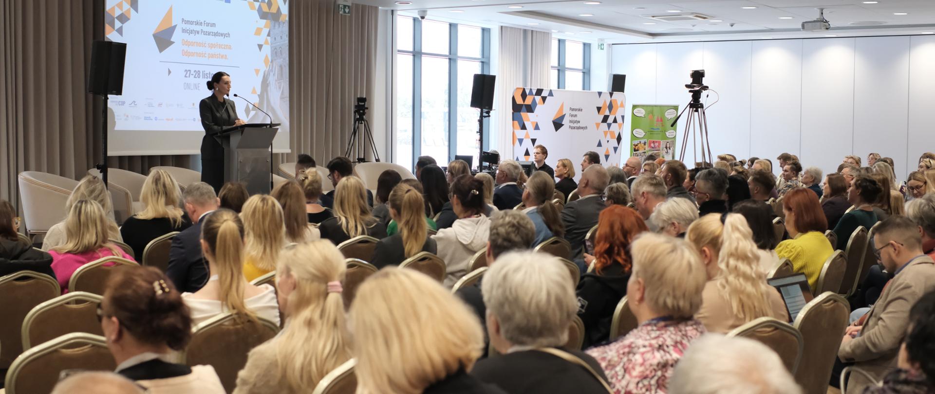 Na pierwszym planie jest tłum ludzi (słuchaczy). W tle widać kobietę na mównicy (wojewodę).