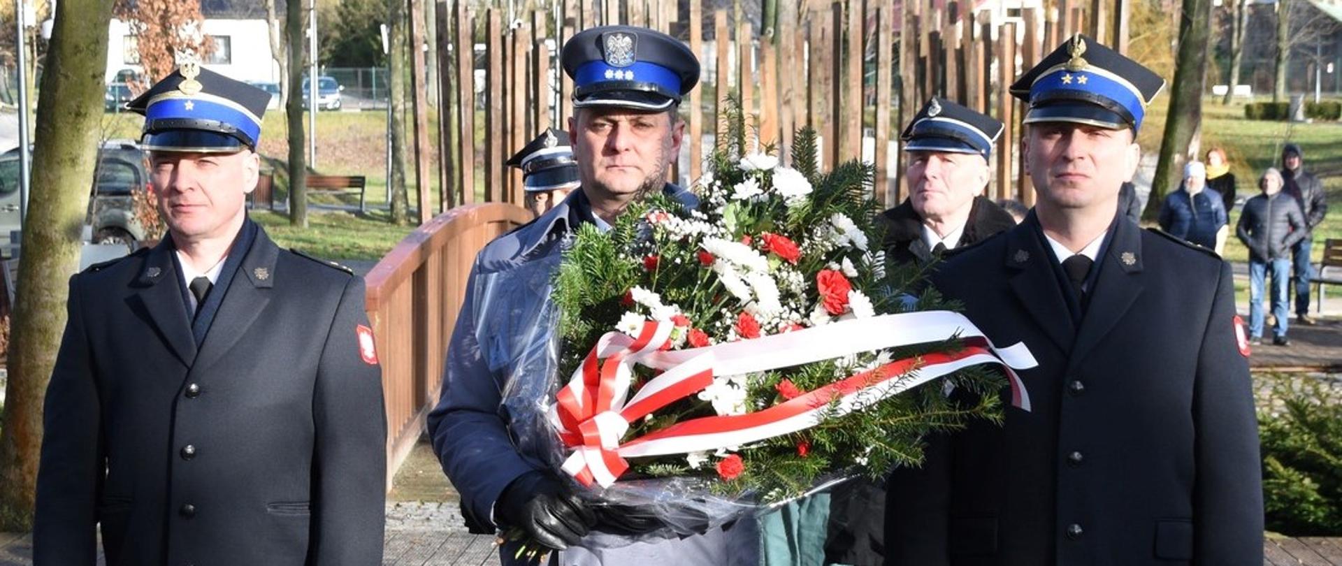 Komendant powiatowy Państwowej Straży Pożarnej w Jędrzejowie mł. bryg. Mariusz Stolarczyk wraz ze swoim zastępcą bryg. Tomaszem Szymańskim w umundurowaniu wyjściowym w płaszczach. Pomiędzy strażakami stoi policjant także w umundurowaniu wyjściowym trzymając w rękach wiązankę. 