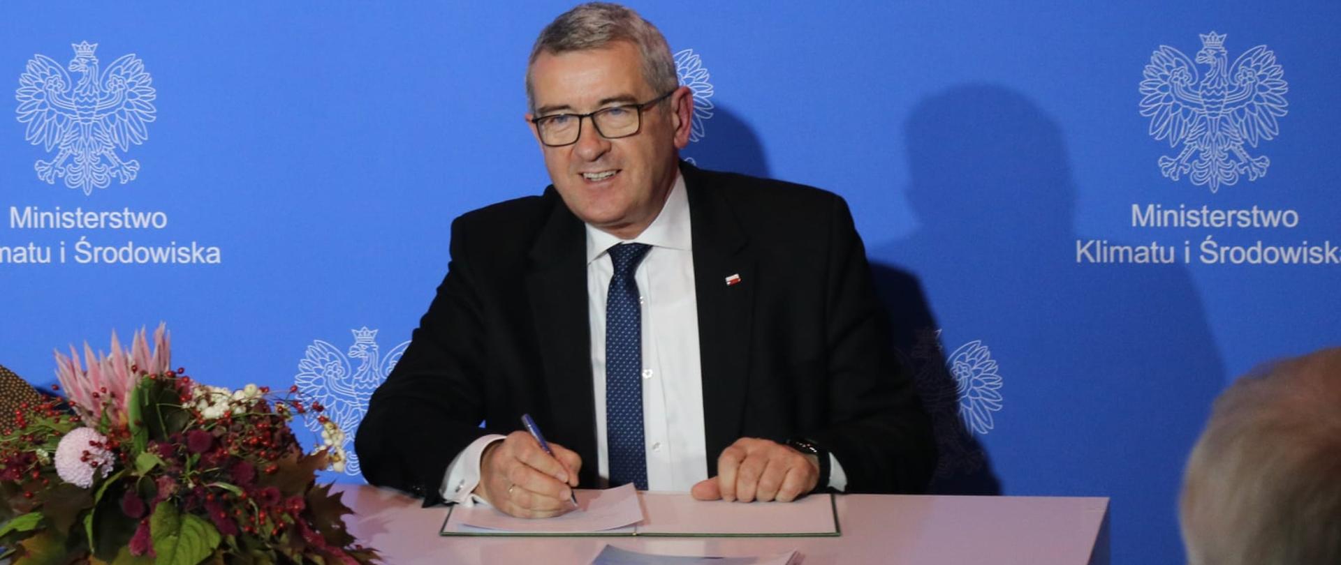 „Porozumienie sektorowe rzecz rozwoju gospodarki wodorowej w Polsce” podpisane – uroczystość z udziałem wiceministra Wojciecha Murdzka, wiceminister Wojciech Murdzek podpisuje „Porozumienie sektorowe rzecz rozwoju gospodarki wodorowej w Polsce”, siedzi przy stole, za nim baner Ministerstwa Klimatu i Środowiska.