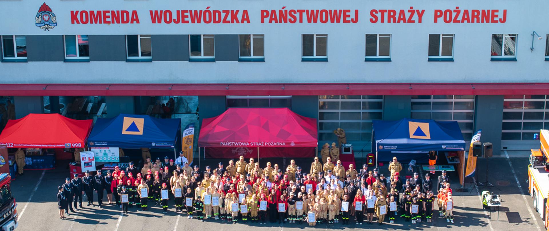 Szeroki widok z góry na elewację budynku Komendy Wojewódzkiej PSP w Opolu. Biały budynek z czerwonymi akcentami i rzędami okien. Na górze duży czerwony napis: „KOMENDA WOJEWÓDZKA PAŃSTWOWEJ STRAŻY POŻARNEJ”. Przed budynkiem, na placu, stoi bardzo duża grupa ludzi – kilkaset osób – ustawionych do wspólnego zdjęcia. Wszyscy w różnych mundurach strażackich (dorośli i dzieci, w tym w mundurach młodzieżowych). Przed grupą, pośrodku, widać dwa czerwone namioty. Lewy namiot z numerem alarmowym 112, prawy z napisem na niebieskim banerze. Plac jest pusty przed grupą, widać tylko cienie. W tle u góry, na dachu, widać instalację fotowoltaiczną.