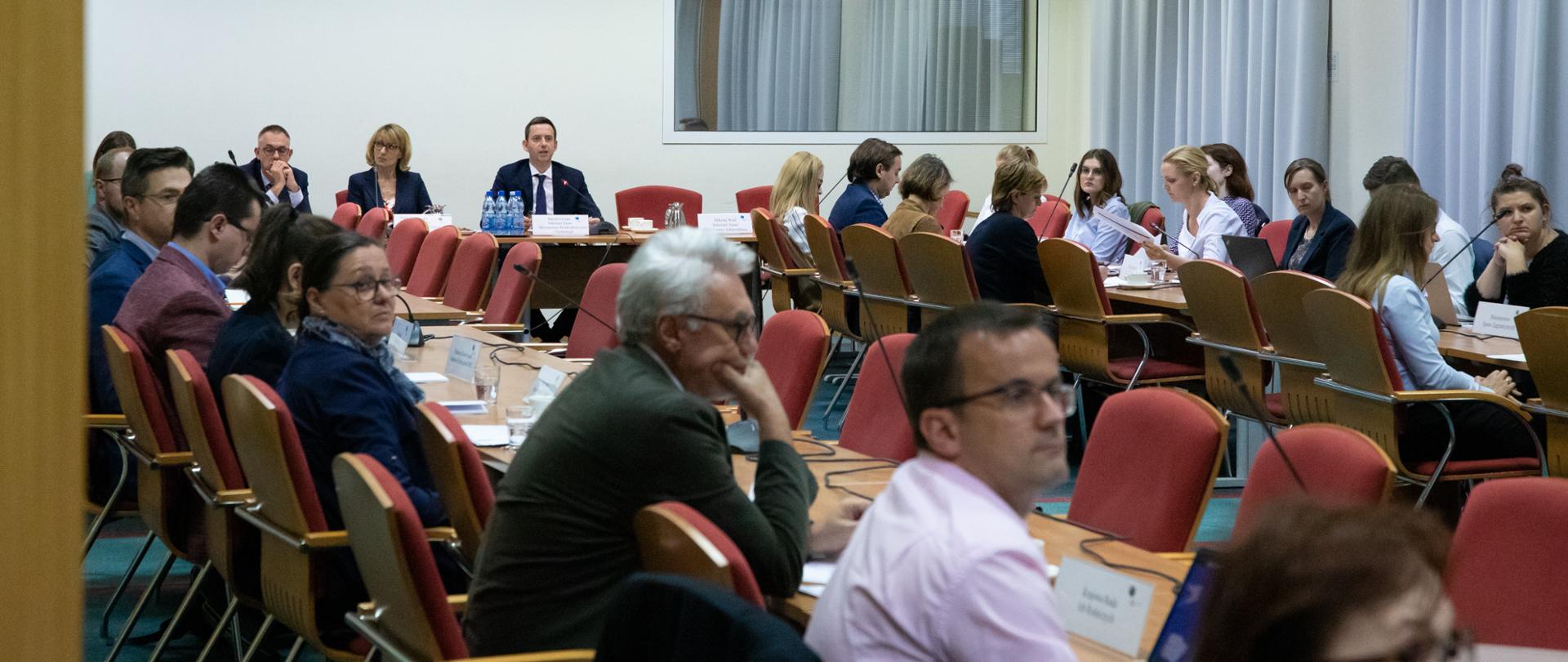 Widzimy uczestników spotkania siedzących wzdłuż stołów.