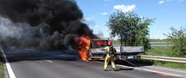 Zdjęcie przedstawia pożar samochodu dostawczego. Pożarem objęta jest kabina samochodu . Widać czarny dym oraz strażaka, który gasi pożar. Auto stoi na poboczu drogi. 