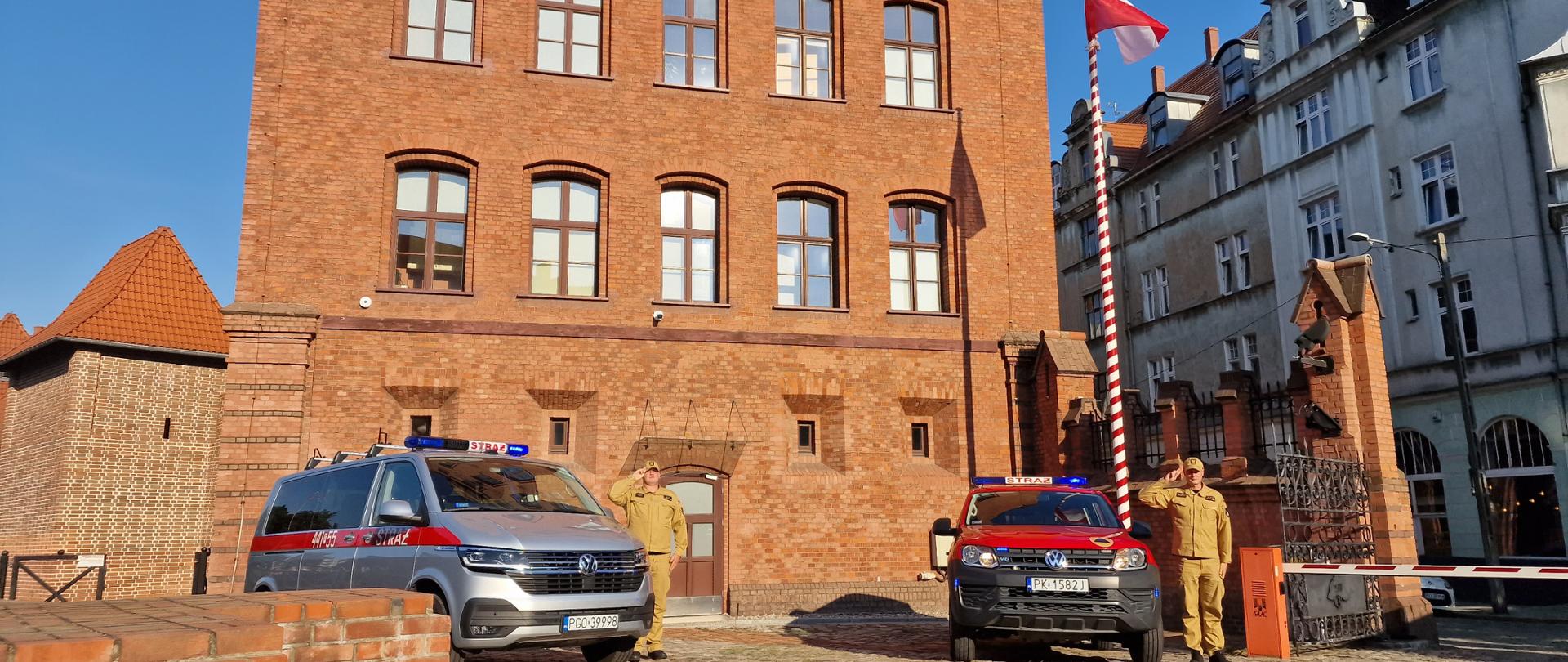 Dwaj strażacy stojący przy samochodach strażackich z włączonymi sygnałami oddają honory. W tle strażnica Komendy Wojewódzkiej PSP w Poznaniu oraz powiewająca na maszcie biało-czerwona flaga