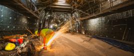 Pracownik w odzieży ochronnej tnie metal przy użyciu szlifierki kątowej wewnątrz budowanego tunelu; jasne iskry rozświetlają wnętrze konstrukcji z rusztowaniami i zbrojeniem.
