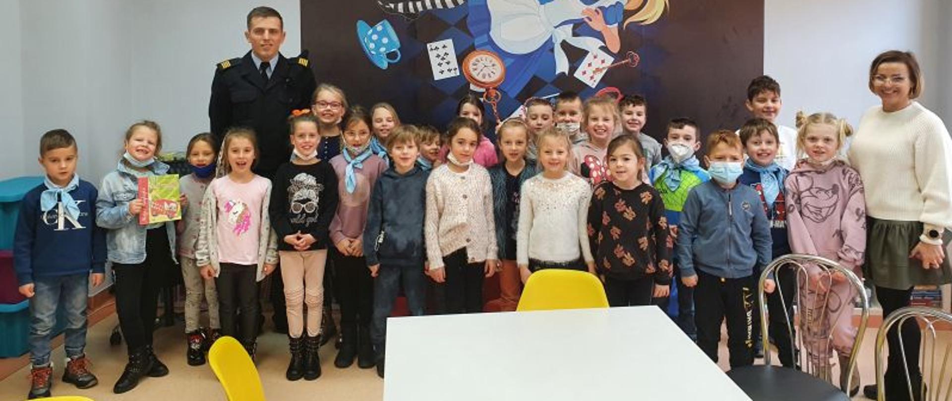Zdjęcie grupowe. Wszystkie dzieci, wychowawca grupy oraz strażak stoją i pozują do zdjęcia w pomieszczeniu czytelni. W tle zdjęcia na ścianie przedstawiające bajkę Kopciuszek