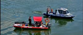 Widok z boku. Łódź specjalistycznej grupy sonarowej Państwowej Straży Pożarnej wraz z łodzią Policji na tafli jeziora. Funkcjonariusz policji wychyla się w oknie łodzi aby wymienić informacje ze strażakami. 