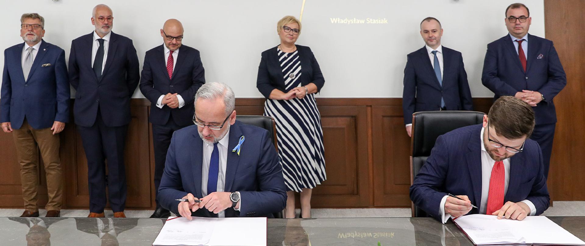 Przy stole siedzą wiceminister Andrzej Śliwka i minister Adam Niedzielski, podpisują dokumenty. W tle inni uczestnicy spotkania. 