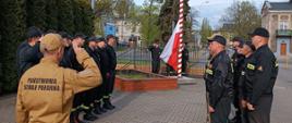 Na zdjęciu strażacy z Jednostki Ratowniczo-Gaśniczej w KP PSP w Pabianicach podczas zmiany służby z okazji Dnia Flagi Rzeczypospolitej Polskiej. Na pierwszym planie Dowódca Jednostki dokonujący zmiany służby, w tle poczet flagowy dokonujący podniesienia flagi na maszt. Strażacy występują w umundurowaniu służbowym (piaskowym) oraz koszarowym (czarne).