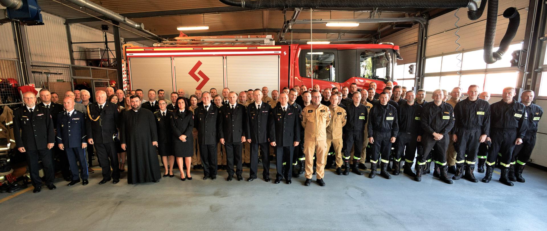 Grupa strażaków w mundurach stoi rzędem w dużym garażu, przed wozem strażackim. Wśród nich jest również ksiądz w czarnej sutannie oraz kilka osób w cywilnych ubraniach.