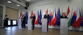 Ministrowie finansów państw Grupy Wyszehradzkiej stoją przed mównicami na tle flag