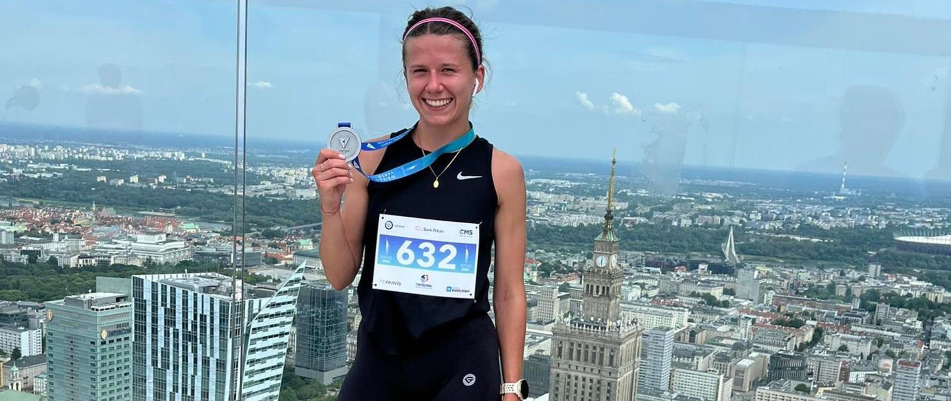 Zdjęcie pamiątkowe z tarasu widokowego budynku Barso Tower. Na pierwszy planie ubrana w sportowy strój kobieta. W ręku trzyma pamiątkowy medal, na koszulce numer startowy w tle rozciągająca się po horyzont panorama Warszawy. 